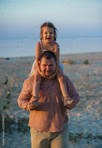 A father and his little daughter play and dance happily on a sandy lake beach at sunset. Happy childhood. The father put his daughter on his neck.