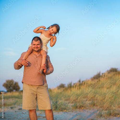 A father and his little daughter play and dance happily on a sandy lake beach at sunset. Happy childhood. The father put his daughter on his neck.