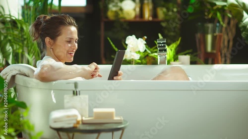 A middle-aged woman enjoys a luxurious bubble bath, leisurely scrolling on her tablet amidst lush greenery, blending modern connectivity with tranquil self-care.