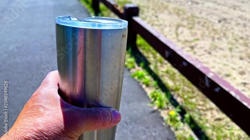 First-person perspective of a man's hand holding a reusable stainless steel travel mug while walking on a path in a park on a sunny day