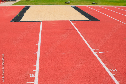 Φωτογραφία Red athletics runway with white distance markings leading toward a long jump sandpit bordered by black rubber and grass