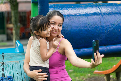 Young Latina mother taking a selfie with her daughter while hugging and smiling at playground
