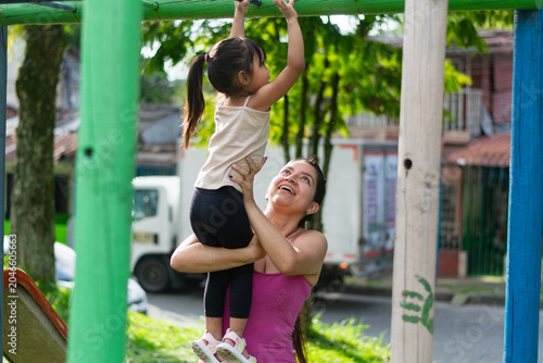 Canvas Print Smiling Latina mother lifting her daughter to help her cross the monkey bars at