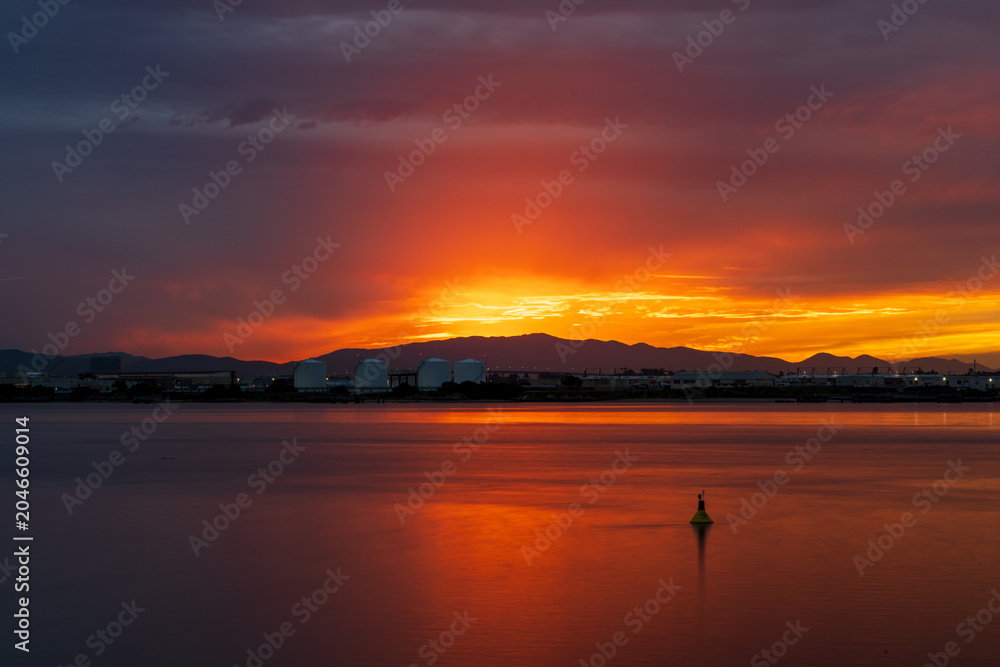 Fototapeta premium Fiery Sunset Over Industrial Waterfront with Calm Bay Water
