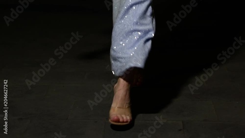 Horizontal night city street video. 60 fps. White woman in long blue shiny floor-length dress with bright rhinestones, paillette, sparkles, sequins walks on sidewalk. Female legs in sandals