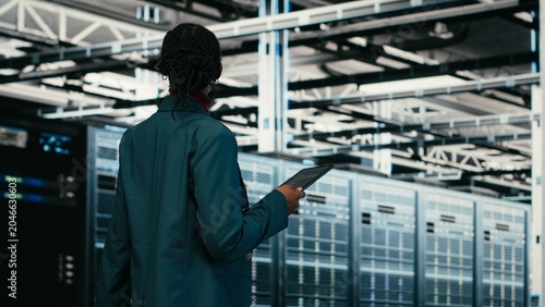Tableau sur toile Female programmer walking in data center using tablet to write code, compiling software patches, African American woman in server hub using device application to run programming script, camera B