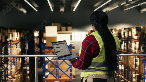 Fotografi African American worker using laptop on elevated platform in fulfillment center, wearing high visibility safety vest, Employee manages logistics, tracking parcels and coordinating order fulfillment