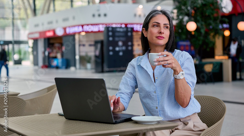 Professional woman enjoying a coffee break while focusing on her work using a laptop at a contemporary cafe, embodying remote productivity and flexible work life balance