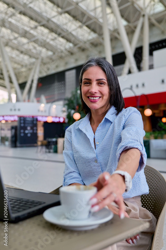 Smiling businesswoman sitting at an airport cafe table with a laptop, reaching for a cup of coffee, symbolizing modern work life balance and digital nomad lifestyle while traveling