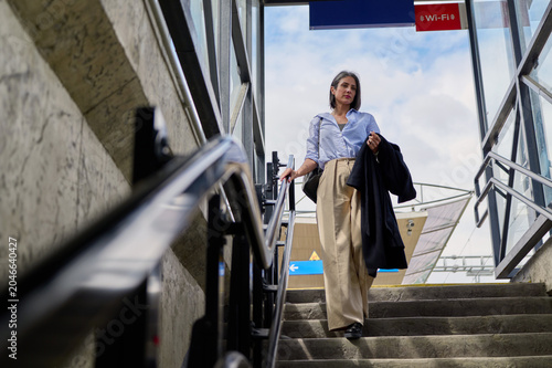 Businesswoman walking down stairs at a modern urban transit station, holding an overcoat and bag, representing daily commute, city life, and professional travel