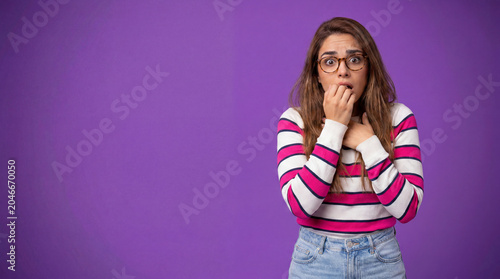 A young woman with glasses and a striped sweater bites her nails, expressing fear or anxiety against a purple background
