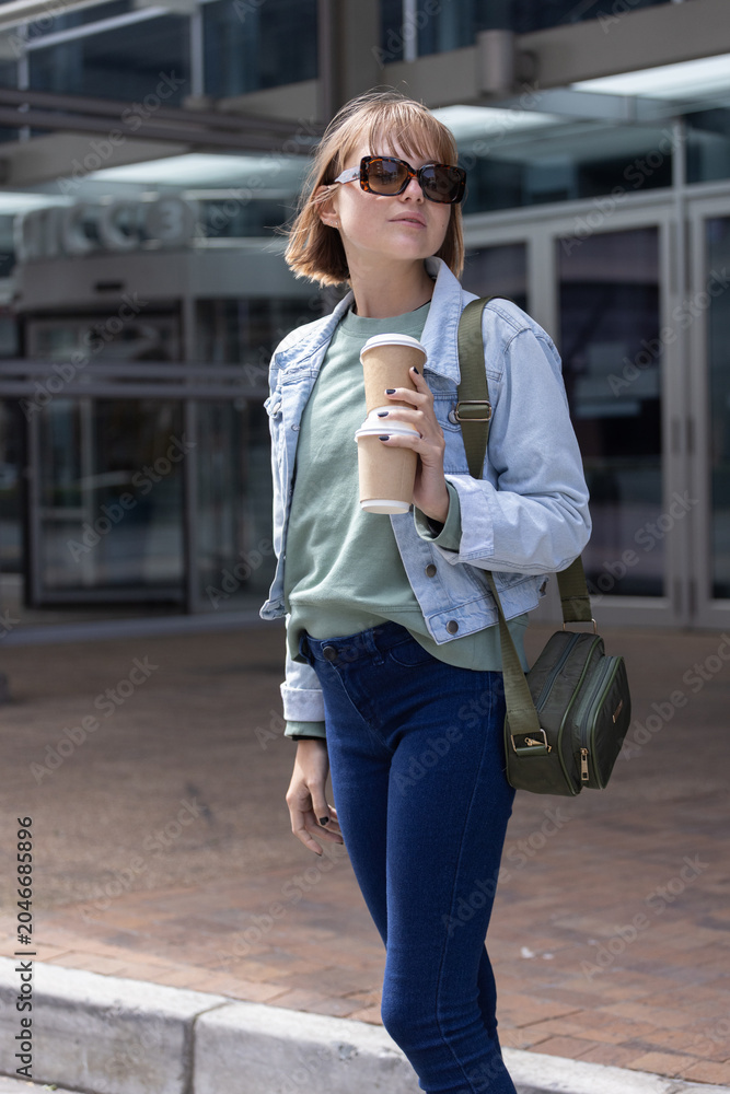 Fototapeta premium Female standing on brick curb at glass entrance wearing denim jacket holding stacked takeaway cups