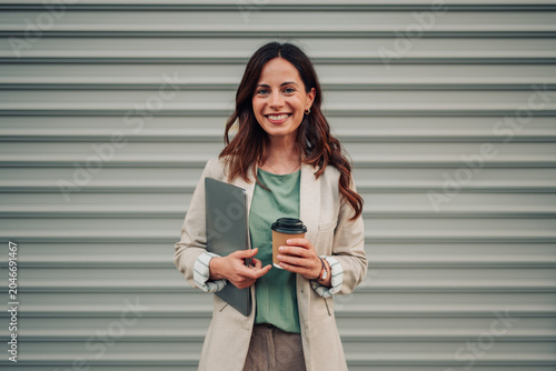 Fotografie Young businesswoman smiling, holding a laptop and take away coffee cup, standing