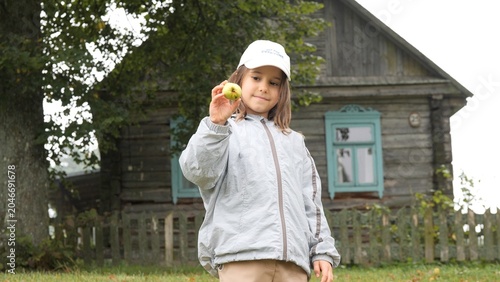 Canvas-taulu Young girl holding a fresh apple in front of a rustic wooden house, smiling outd