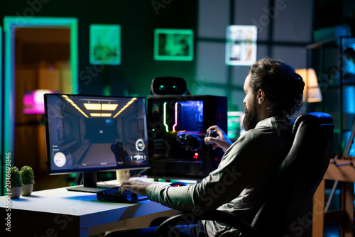 Fotografi Man in dark living room playing videogame on gaming PC at computer desk, relaxing by shooting enemies, putting on headphones