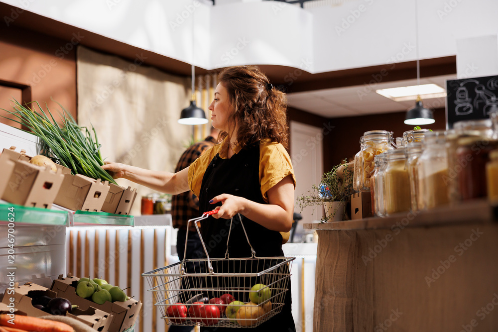Obraz premium Woman in low carbon footprint zero waste store interested in purchasing food with high nutritional value. Client fills shopping basket with pesticides free produce in local neighborhood shop