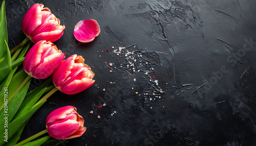 Four vibrant pink tulips with green aloe vera leaves arranged on a textured dark background, romantic spring floral arrangement