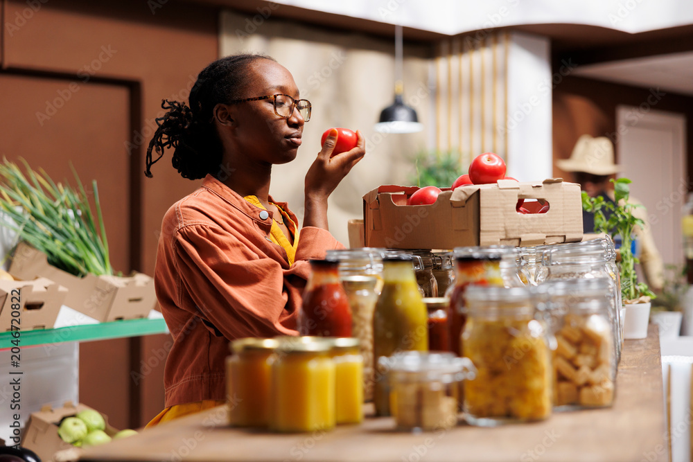 Obraz premium Black woman shops at local market, supporting local producers and choosing eco friendly products like fresh fruits and vegetables. African American lady near the glass checking out the tomatoes.