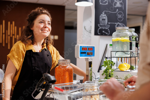 Fotografija Cheerful woman at checkout counter in zero waste shop selling healthy lentils in jar to customer with green living lifestyle