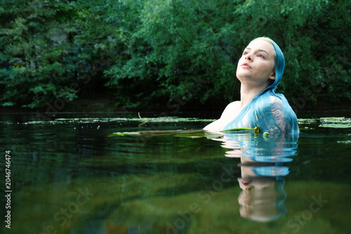 A blue haired woman emerges from a tranquil forest pond while reflections water plants and dark green surroundings create a mystical atmosphere of connection with nature