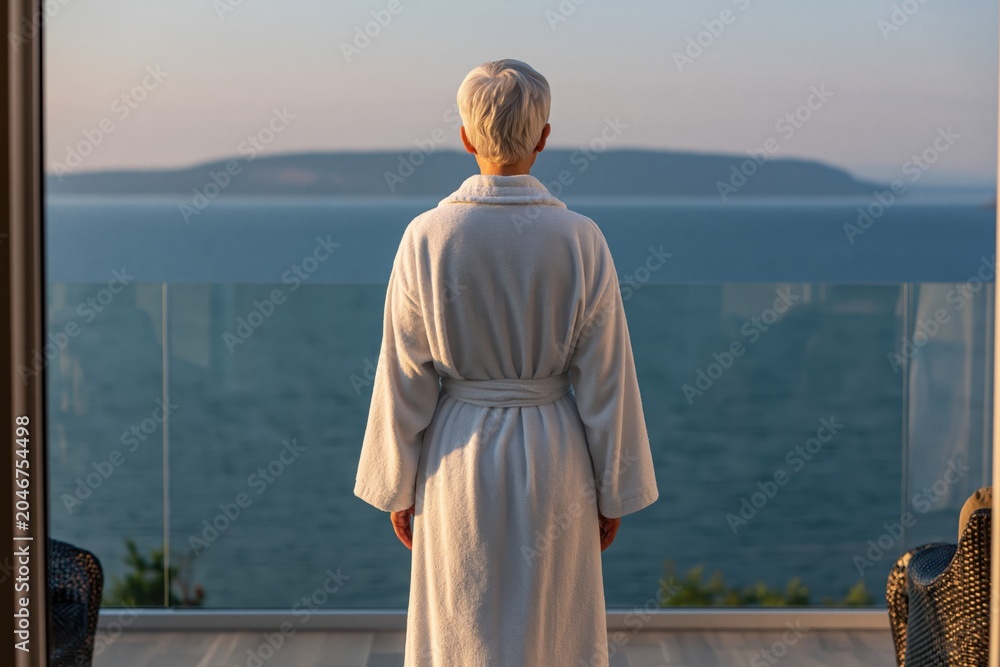 Fototapeta premium Woman senior in bathrobe on balcony gazing at tranquil ocean sea view sunset