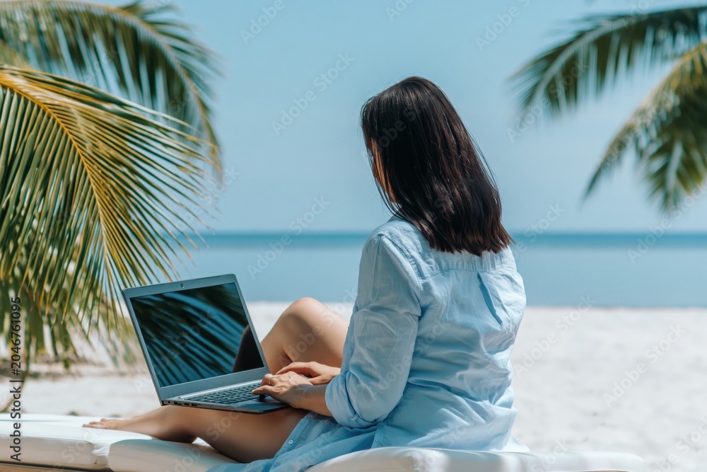 Fototapeta premium Woman working on laptop on tropical beach, remote digital nomad lifestyle by ocean