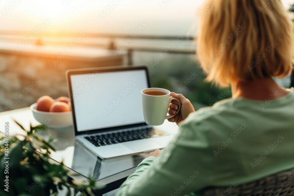 Fototapeta premium Woman working on laptop on balcony at sunset with coffee remote lifestyle evening
