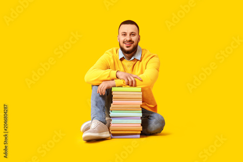 Young man with stack of books sitting on yellow background