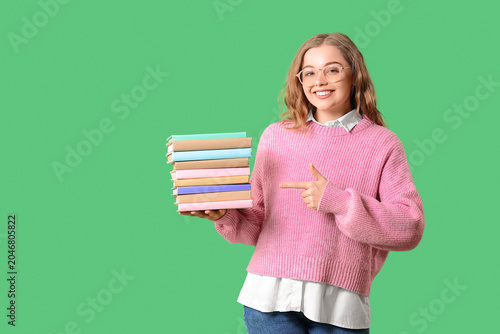 Young woman in eyeglasses pointing at stack of books on green background