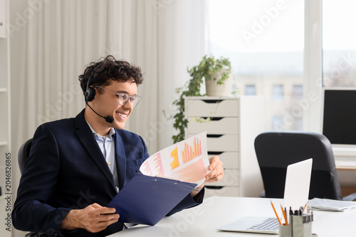 Young male online business consultant with headset, clipboard and laptop vide...