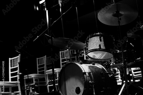 Black and white photograph of a jazz drum set positioned on an empty concert stage surrounded by chairs and microphone stands. Strong contrast, dramatic lighting and large copy space create