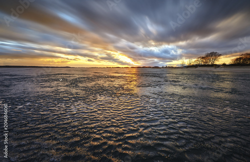 View of the frozen surface of Lake Tarnobrzeg with rippled ice under a dramatic sunset sky with moving clouds Tarnobrzeg, Podkarpackie, Poland.