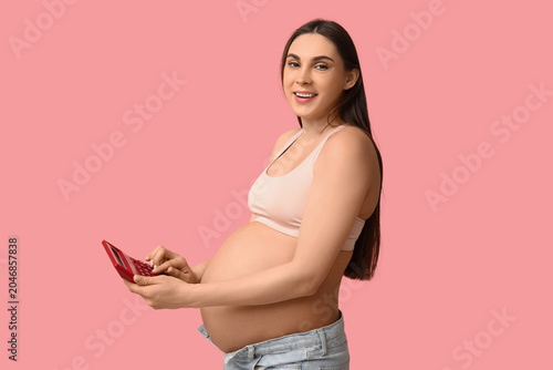 Pregnant young woman with calculator on pink background