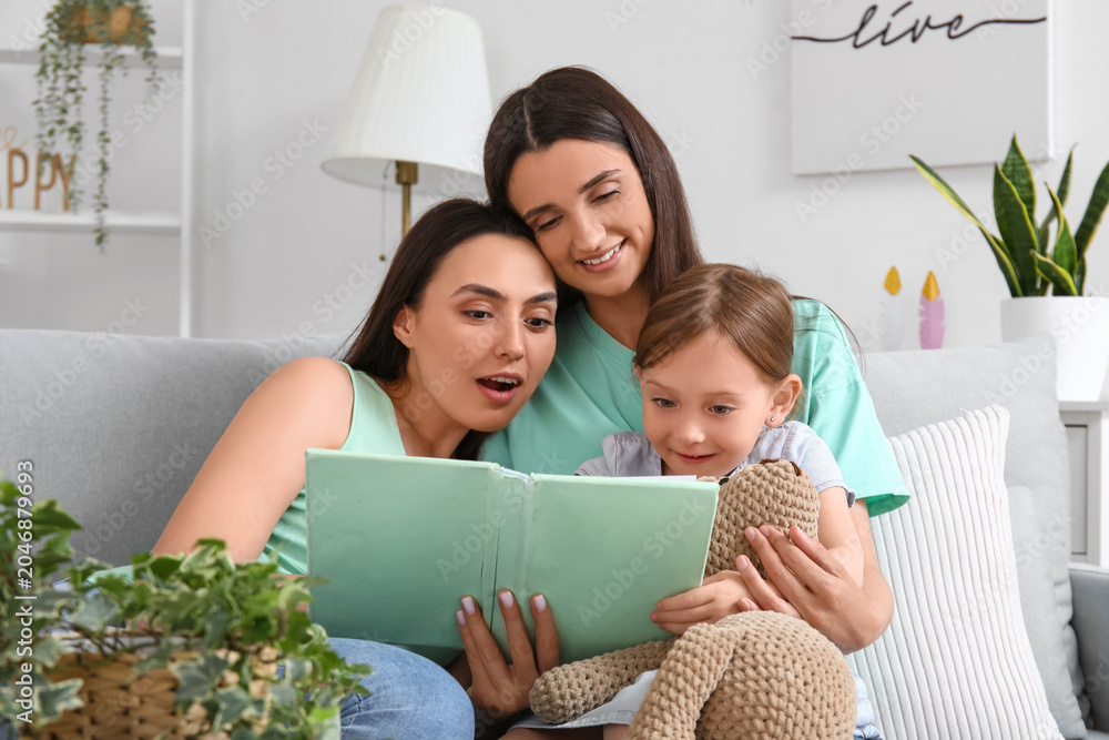 Naklejka premium Young lesbian couple with adopted little girl reading book at home