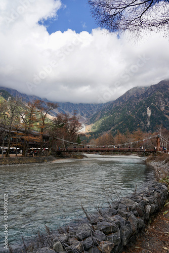 Autumn Mountain Village With Suspension Bridge Over River and Tourists Under Cloudy Sky