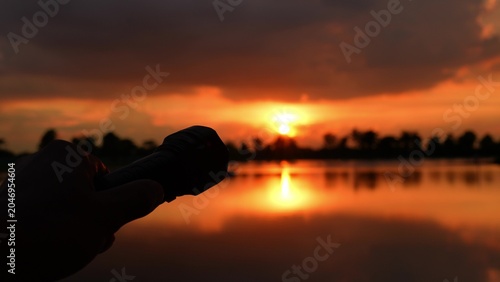 Schilderij op canvas Shining a flashlight toward the sunset sky symbolizes seeking new directions, the search for clarity, guiding one’s path, blending human ingenuity with nature, and the power of hope