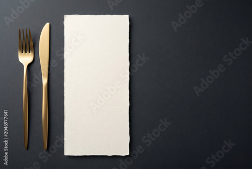 Elegant gold cutlery with blank menu card on dark background. Luxury restaurant table setting with golden fork and knife. Minimalist food stationery mockup for wedding or fine dining.