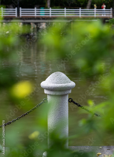 Foto Sturdy gray stone post in focus with rounded dome cap, dark metal chains on sides, calm water and white railing in soft bokeh background, framed by green leaves, dreamy impressionist quality