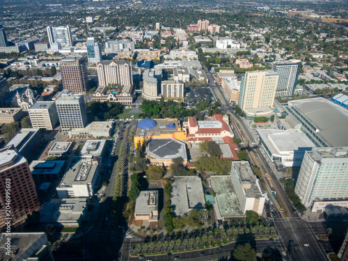 This image presents a high-angle aerial view of a downtown San Jose on a clear day. The scene captures a mix of modern office towers, hotels, civic buildings, and commercial structures arranged along 