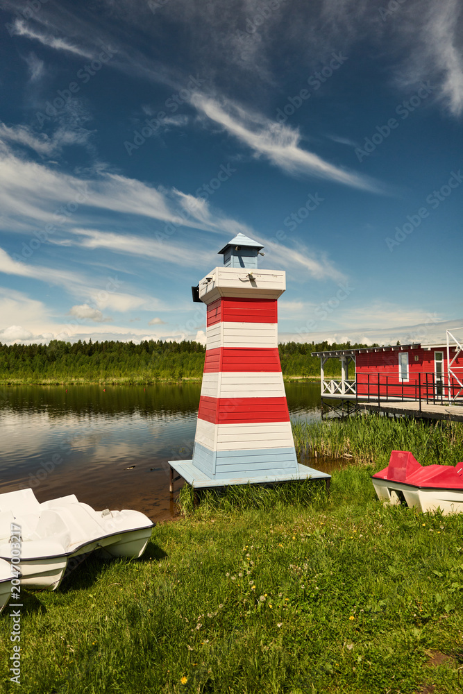 Obraz premium A red and white striped wooden lighthouse stands on grassy lake shore next to small pedal boats and a red boathouse. The scene shows calm reflective water, a