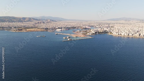 Aerial drone view flying over the calm aegean sea, revealing the sprawling urban cityscape and the bustling port of piraeus in athens, greece, with mountains visible in the hazy distance