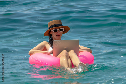 Bild auf Leinwand Remote work digital nomad swimming woman using laptop floating on inflatable ring in blue ocean water during summer vacation