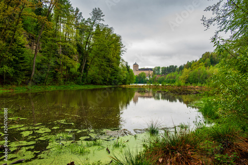 Kost Castle rises above a pond in the Bohemian Paradise region of the Czech Republic. Forests surround the landscape under cloudy skies, creating a scenic historic view.