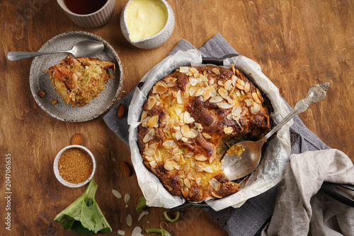 Top view of freshly baked English rhubarb pudding with custard, golden almond crust and a cut slice on plate, served in cast iron skillet on wooden background flat lay
