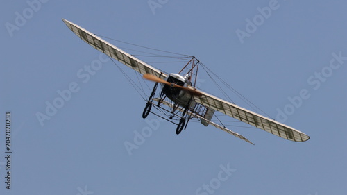 View of a vintage monoplane with a wooden propeller and wire-braced wings flying through a clear blue sky during an airshow in La Ferte-Alais, Ile-de-France, France.
