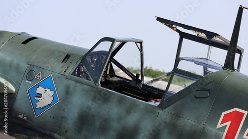 View of the open cockpit and canopy of a vintage Messerschmitt Bf 109 fighter aircraft with a wolf emblem on the fuselage at the airfield in La Ferte-Alais, Ile-de-France, France.