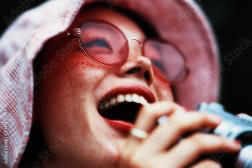 Close-up of a smiling woman with freckles and sunglasses, capturing a joyful moment while holding a camera outdoors.