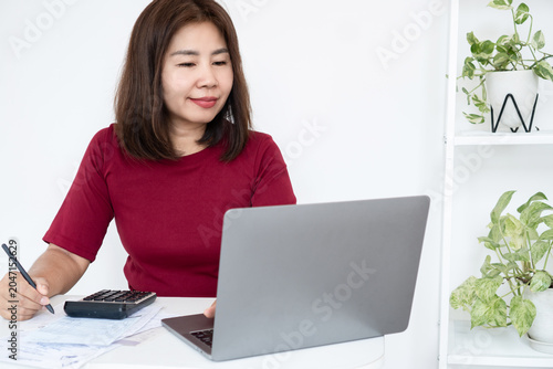 happy Asian businesswoman smiling while working on a laptop at her office desk, holding a pen with a calculator. Concept of financial success, remote work, small business, and happiness