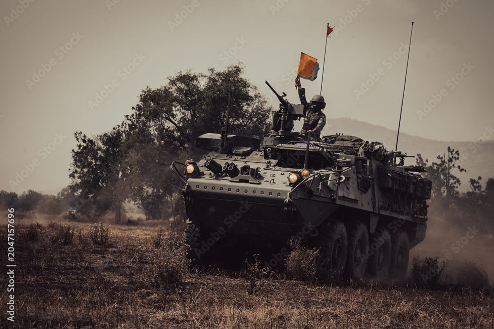 Naklejka premium Dramatic shot of an eight-wheeled green military armored vehicle (APC) advancing through a dense dust and haze cloud on a dry dirt field under heavy golden sunset light.