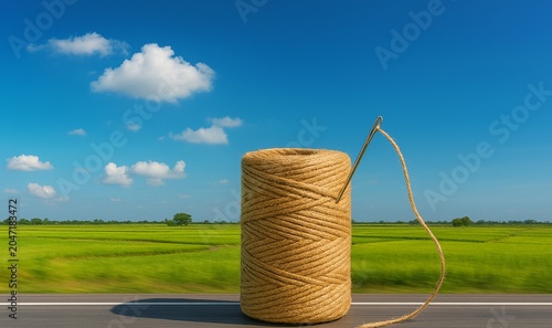 A spool of rustic brown twine with a needle, placed on a road surface against a backdrop of a lush green rice field under a vibrant blue sky.